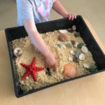 Child engaging with sensory sand experience with seashells and starfish